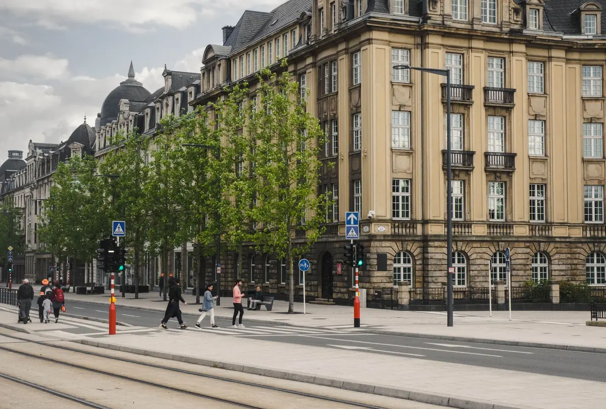 Piesi przechodzą przez jezdnię po przejściu dla pieszych z sygnalizacją i niebieskimi znakami przejścia, na pierwszym planie tory tramwajowe oddzielone od jezdni