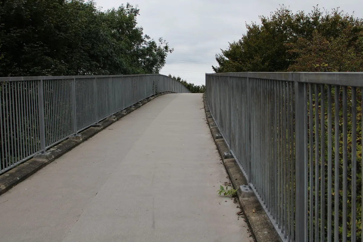 Betonowa kładka dla pieszych wznosząca się łukiem, po obu stronach stalowe ocynkowane balustrady z gęstymi pionowymi szczeblinami