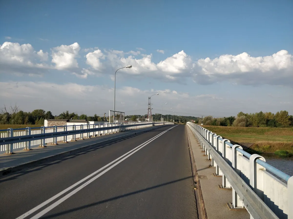 Most drogowy nad rzeką z chodnikami po obu stronach, wzdłuż krawędzi białe słupki z niebieską poręczą i stalową prowadnicą bariery