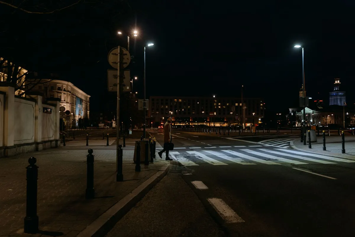 Mężczyzna z torbą przechodzi nocą przez przejście dla pieszych na szerokiej ulicy w centrum miasta, przejście oświetlają latarnie, przy chodniku stoją słupki