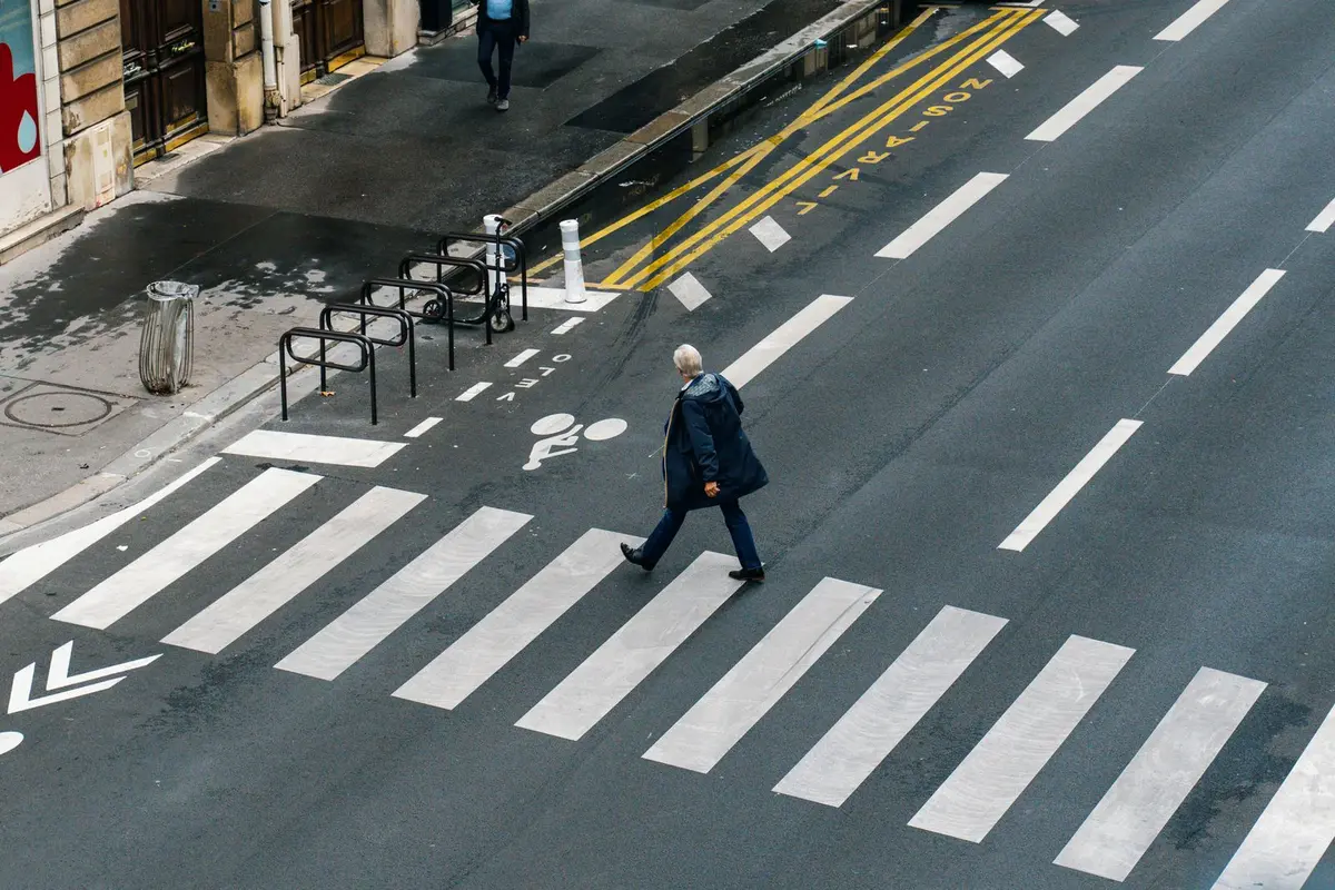 Widok z góry na ulicę w Paryżu: starszy mężczyzna przechodzi przez szerokie przejście dla pieszych, obok pas rowerowy i stojaki na rowery przy chodniku