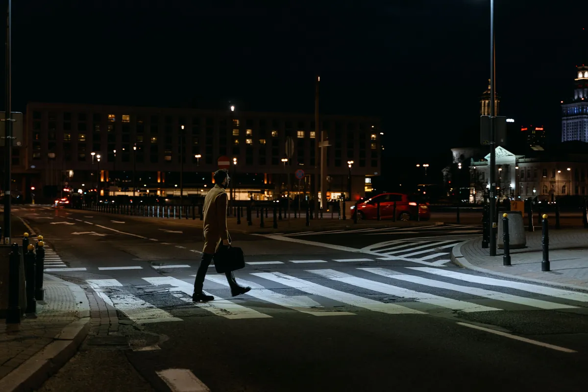 Mężczyzna z teczką przechodzi nocą przez przejście dla pieszych na szerokiej ulicy w mieście, oświetlonej latarniami ustawionymi wzdłuż jezdni
