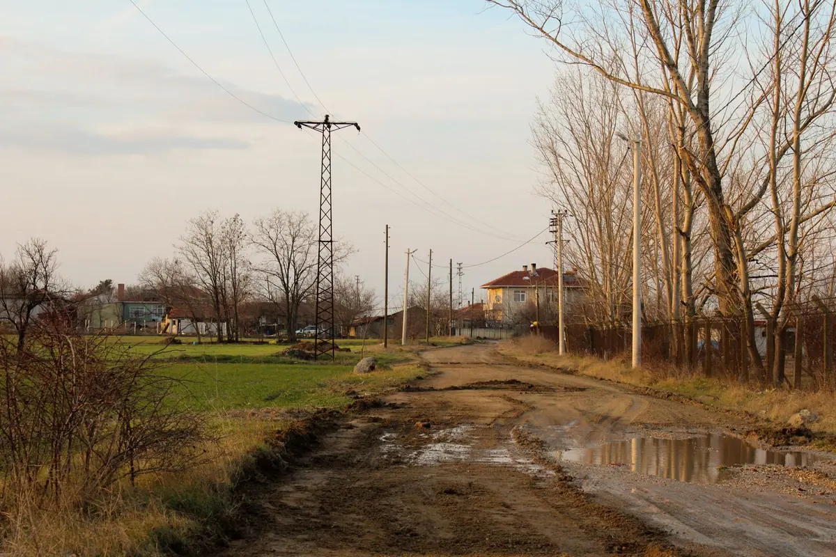 Błotnista droga gruntowa na skraju wsi z kałużami i koleinami, obok słupy energetyczne i domy