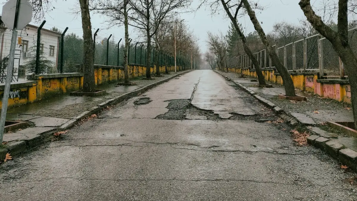 Popękana asfaltowa droga z ubytkami nawierzchni pośrodku jezdni, wzdłuż niej ogrodzenia i nagie drzewa