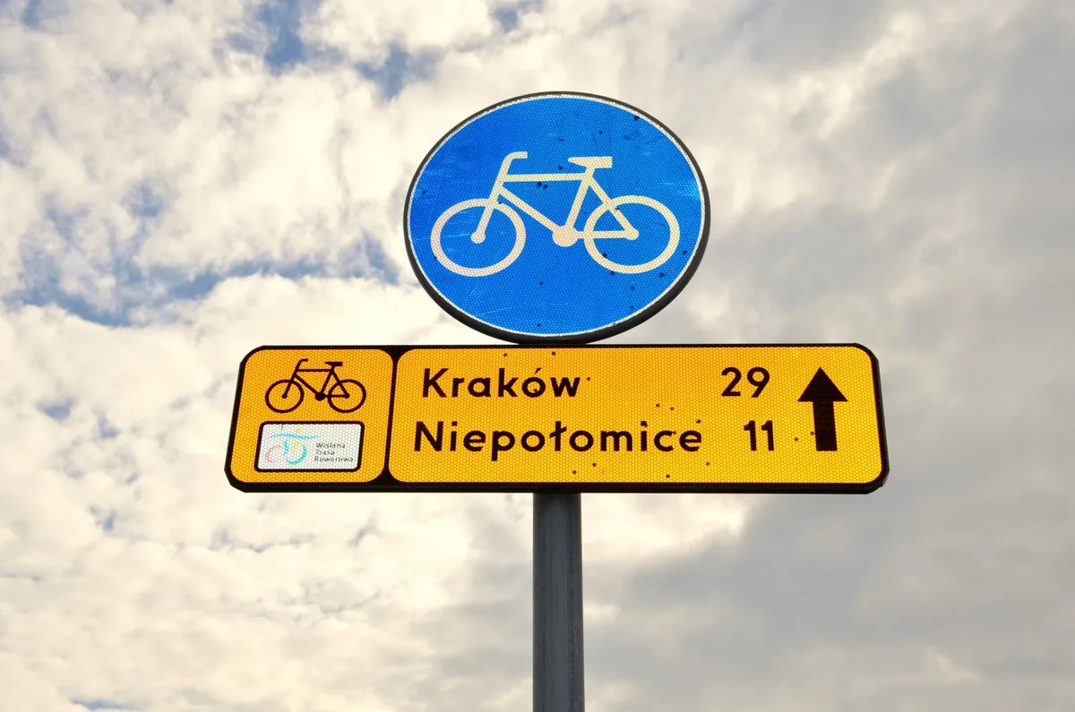 Niebieski okrągły znak z rowerem, pod nim żółta tablica szlaku rowerowego ze strzałką i odległościami do Krakowa (29 km) i Niepołomic (11 km)