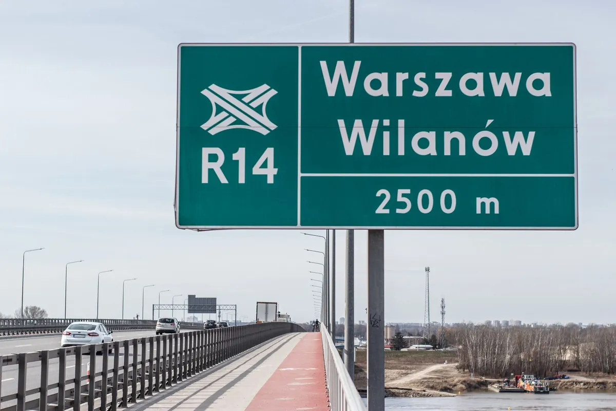Duża zielona tablica nad drogą w Warszawie z symbolem węzła, oznaczeniem R14, napisem Warszawa Wilanów i odległością 2500 m
