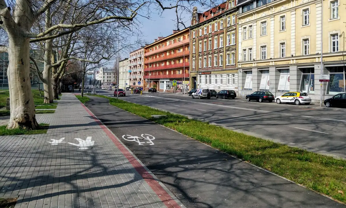 Ulica miejska z chodnikiem z kostki, drogą dla rowerów, pasem trawy i jezdnią, wzdłuż której parkują samochody