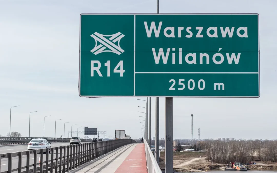 Zielona tablica kierunkowa z napisem Warszawa Wilanów 2500 m na stalowym słupie przy moście, obok bariera i droga dla rowerów