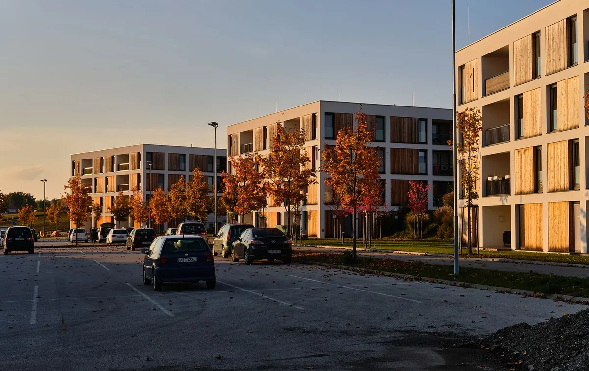 Parking z wyznaczonymi stanowiskami przy czterokondygnacyjnych blokach mieszkalnych, jesienne drzewa i zaparkowane samochody osobowe