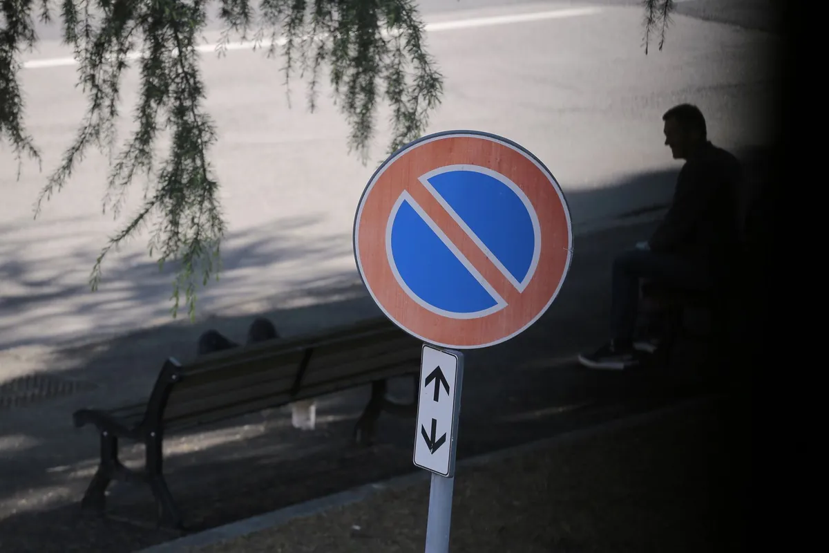 Okrągły znak zakazu postoju z jednym czerwonym pasem i tabliczką ze strzałkami w górę i w dół, w tle ławka w parku