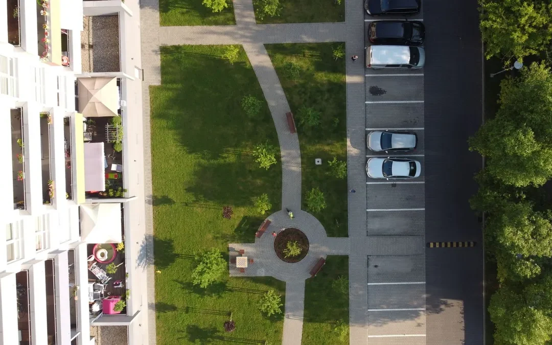 Widok z drona na blok, pas trawnika ze ścieżkami i placykiem oraz parking i jezdnię osiedlową wzdłuż budynku
