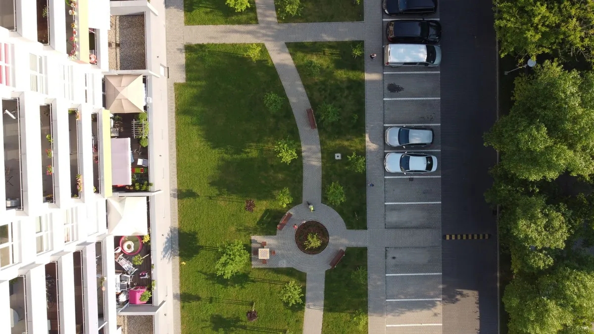 Widok z drona na blok, pas trawnika ze ścieżkami i placykiem oraz parking i jezdnię osiedlową wzdłuż budynku