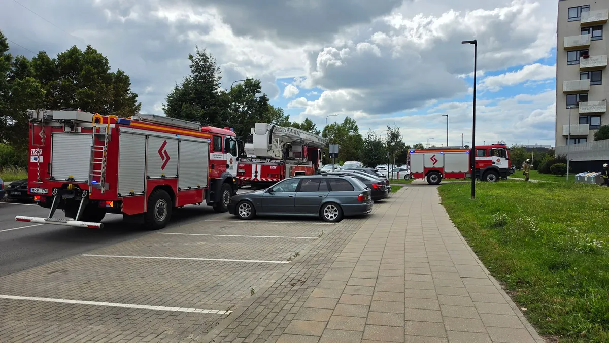 Dwa wozy strażackie, w tym samochód z drabiną mechaniczną, stoją na jezdni przy parkingu osiedlowym obok bloku, trzeci wóz na drodze w głębi