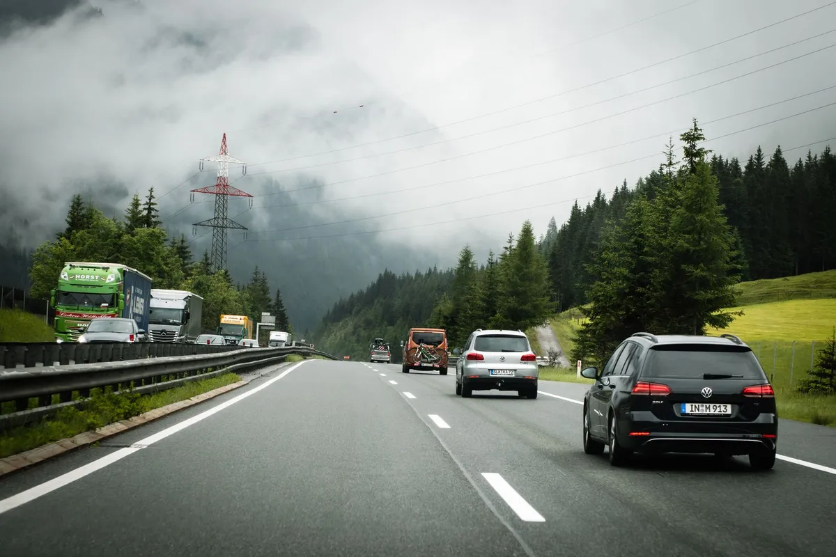 Autostrada w austriackich Alpach w pochmurny dzień: samochody osobowe na jezdni w stronę gór, po drugiej stronie bariery kolumna ciężarówek, w tle zalesione zbocza we mgle