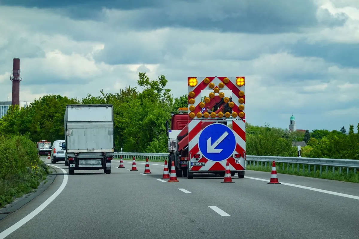 Ciężarówka z tablicą zamykającą w biało-czerwone pasy, z żółtymi lampami i niebieskim znakiem nakazu, stoi na prawym pasie drogi dwujezdniowej za skosem z pachołków
