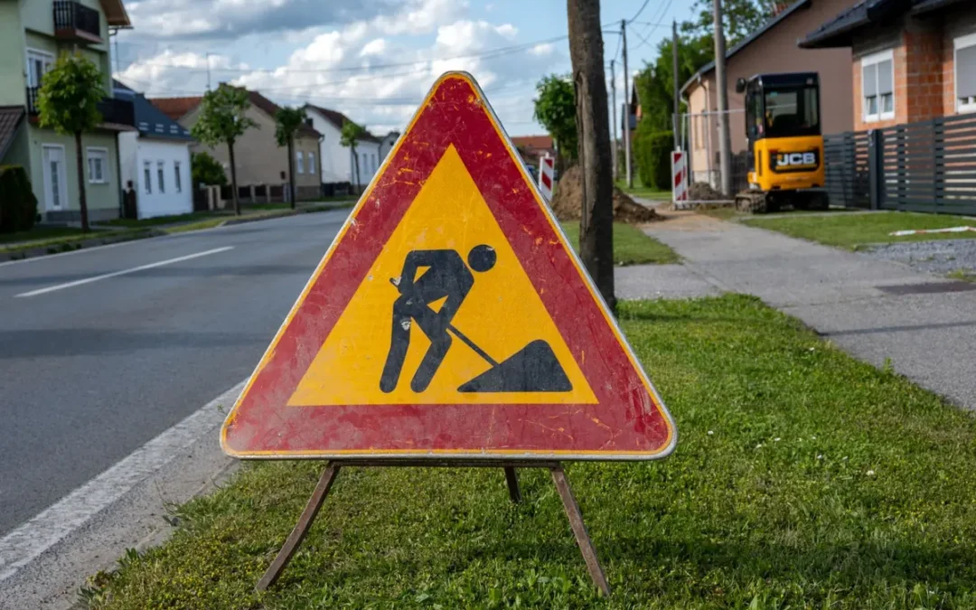 Trójkątny znak roboty na drodze na składanym stojaku, ustawiony na trawniku przy jezdni; w tle koparka i hałda ziemi