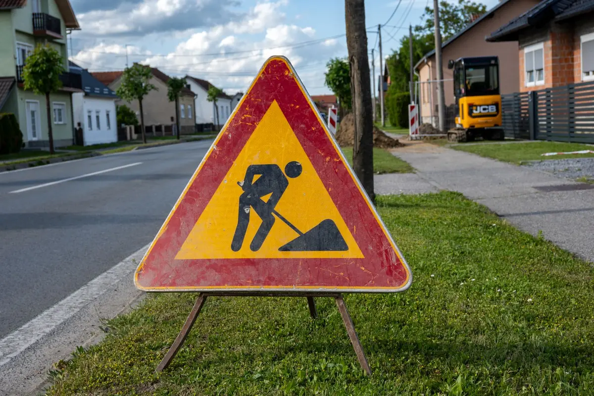 Trójkątny znak roboty na drodze na składanym stojaku, ustawiony na trawniku przy jezdni; w tle koparka i hałda ziemi