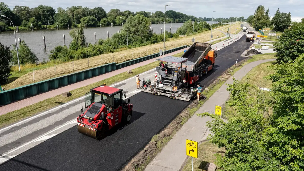 Widok z góry na układanie nowej warstwy asfaltu: rozściełacz, wywrotka i czerwony walec na drodze wzdłuż rzeki