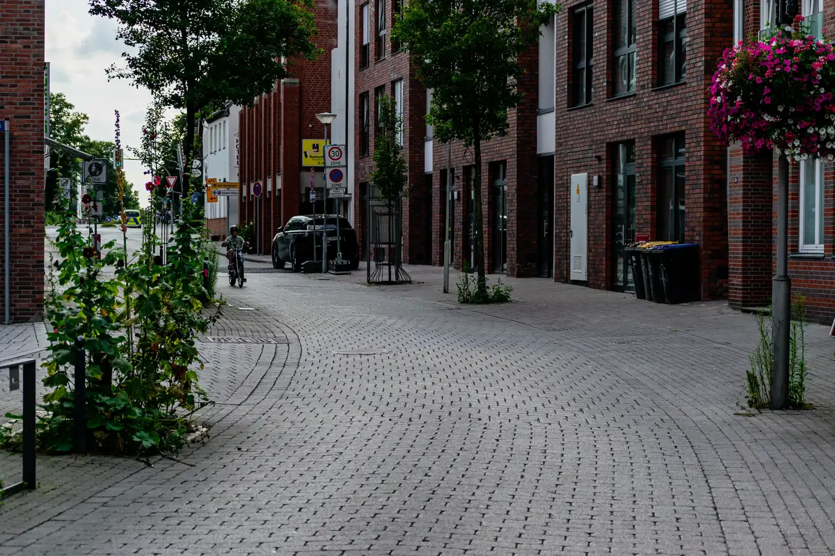 Ulica z jedną nawierzchnią z kostki betonowej bez krawężników, ceglane budynki mieszkalne, w głębi rowerzysta i zaparkowany samochód