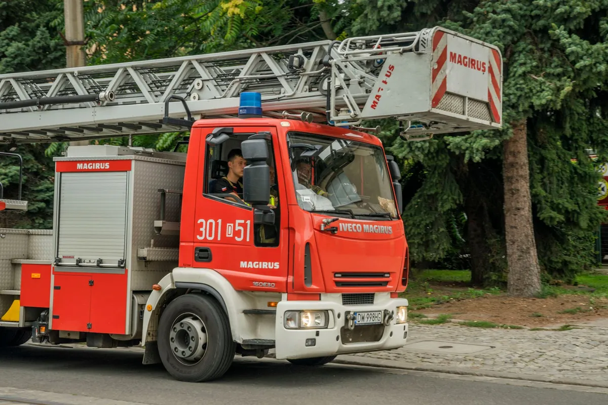 Czerwony samochód straży pożarnej z drabiną mechaniczną Magirus jedzie ulicą wzdłuż drzew