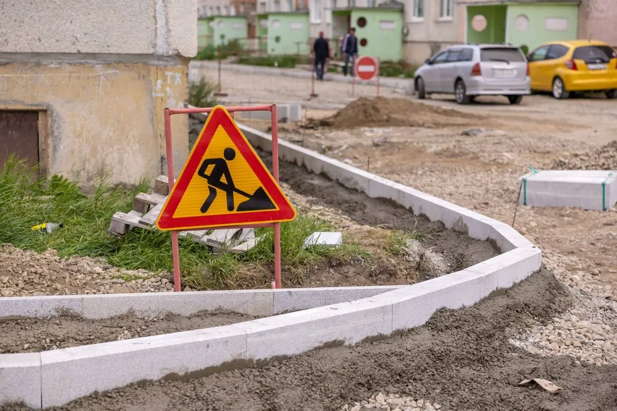 Trójkątny znak roboty drogowe na czerwonym stojaku przy nowo układanych krawężnikach, w tle znak zakazu wjazdu i zaparkowane samochody