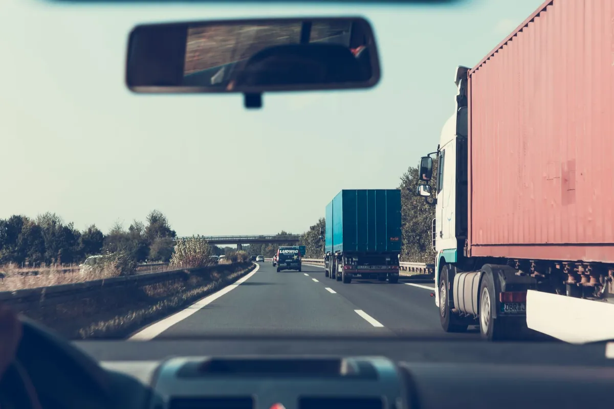 Widok zza kierownicy samochodu osobowego na dwupasmową autostradę, prawym pasem jadą jedna za drugą dwie ciężarówki z naczepami