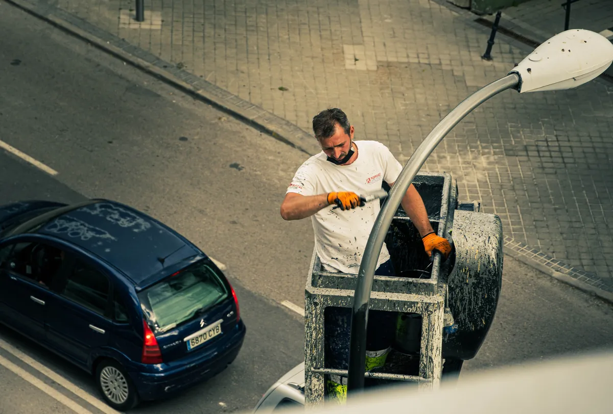 Pracownik w koszu podnośnika przy słupie latarni ulicznej nad jezdnią, obok zaparkowany samochód